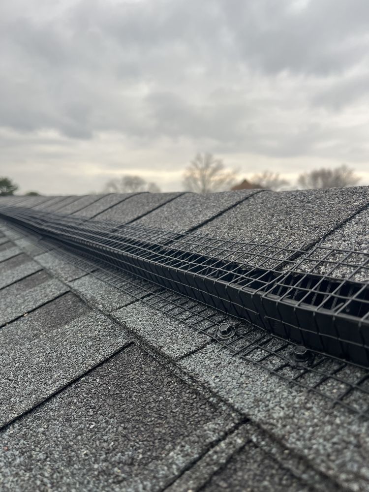 Snow-covered roof shingles under a cloudy sky, with a long black roof vent running along the ridge
