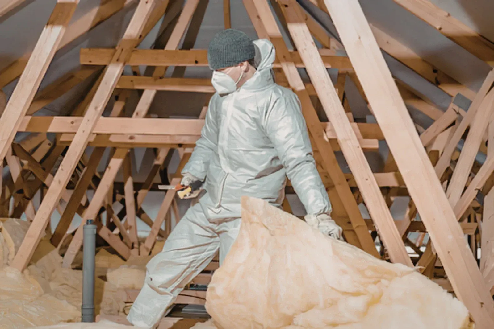 Worker in protective suit insulating an attic, surrounded by wooden roof beams and fiberglass insulation.
