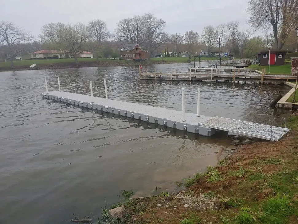 Floating dock on a pond near a grassy shore, with a small red building and bare trees in the background