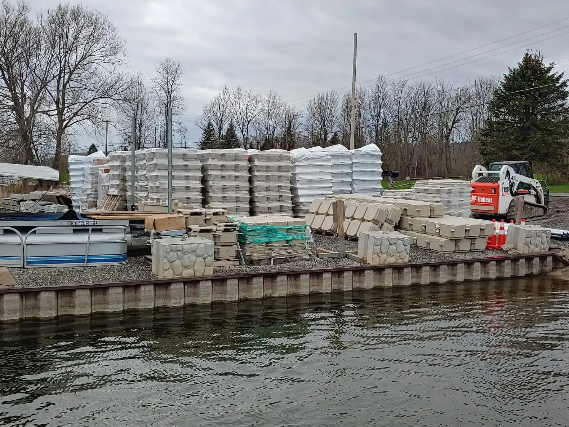 Construction materials stacked beside a waterfront, with a small skid steer on a rocky shoreline under a cloudy sky