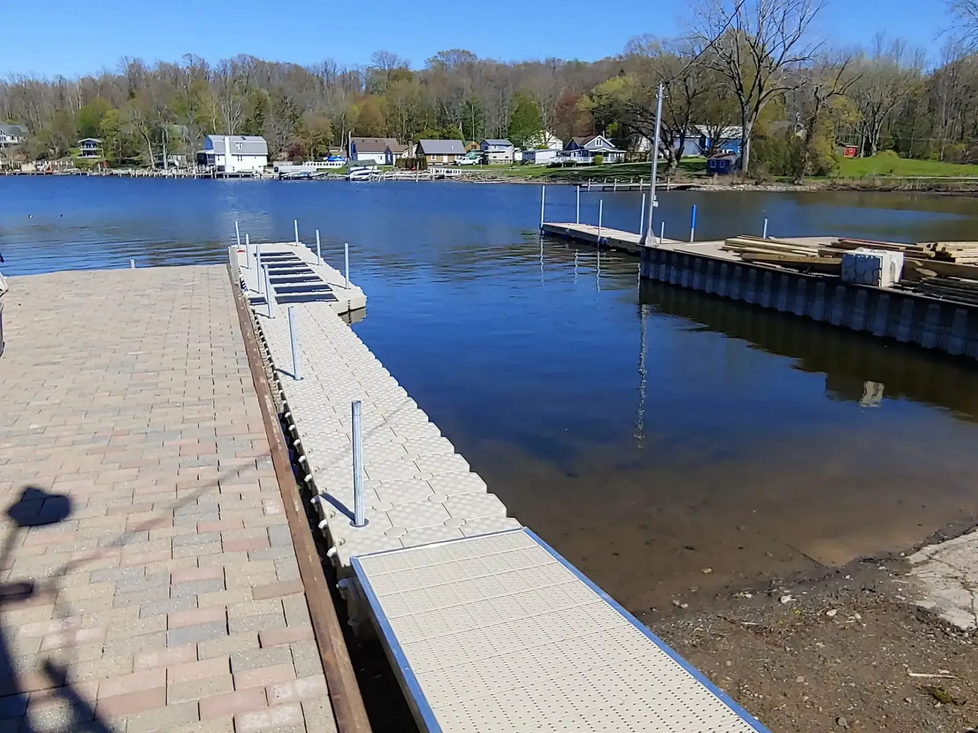 Floating dock on a calm lake beside a paved shoreline, with houses and trees across the water.