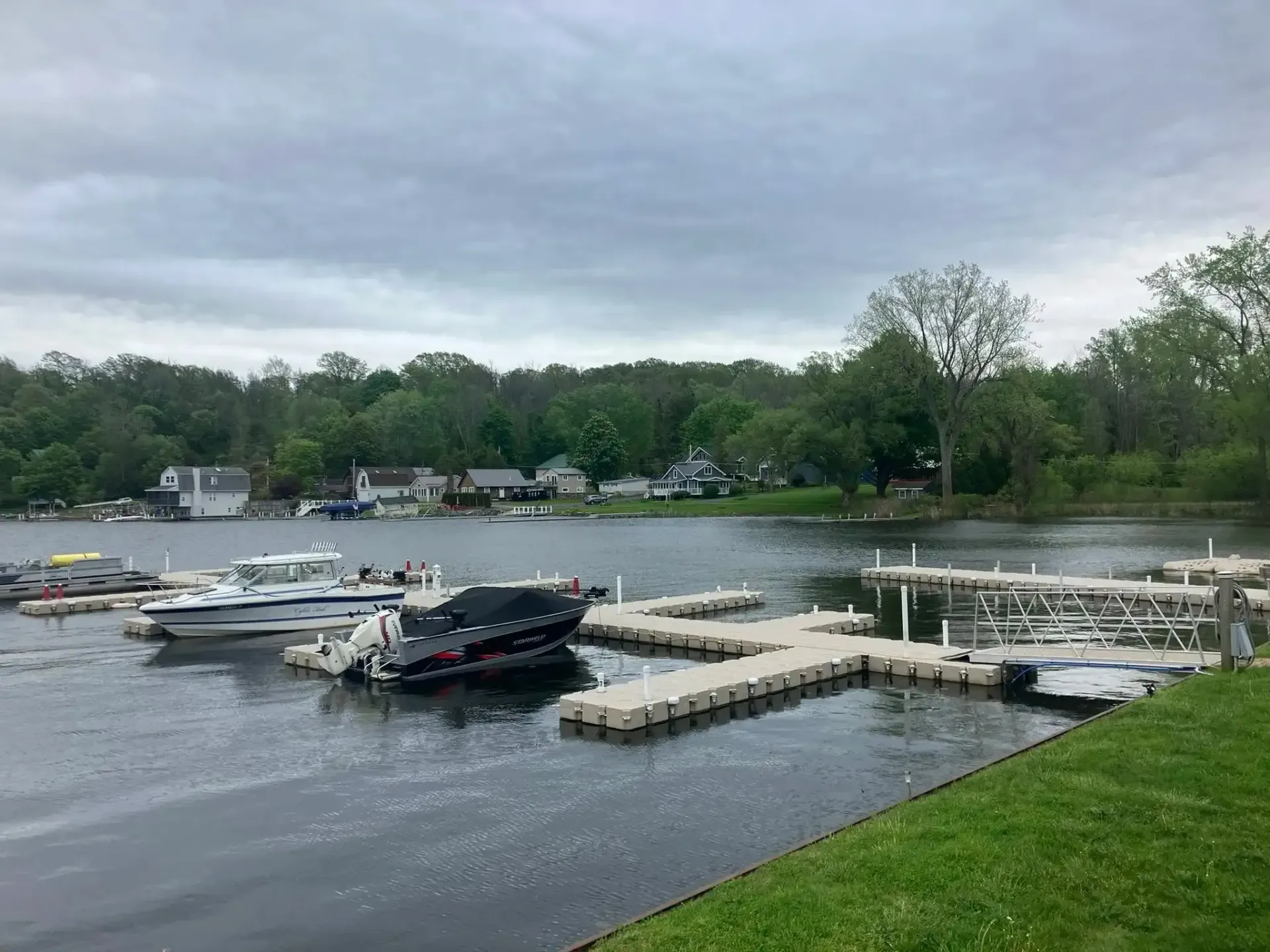 Boats docked at a lakeside marina under a cloudy sky, with trees and grassy shore nearby