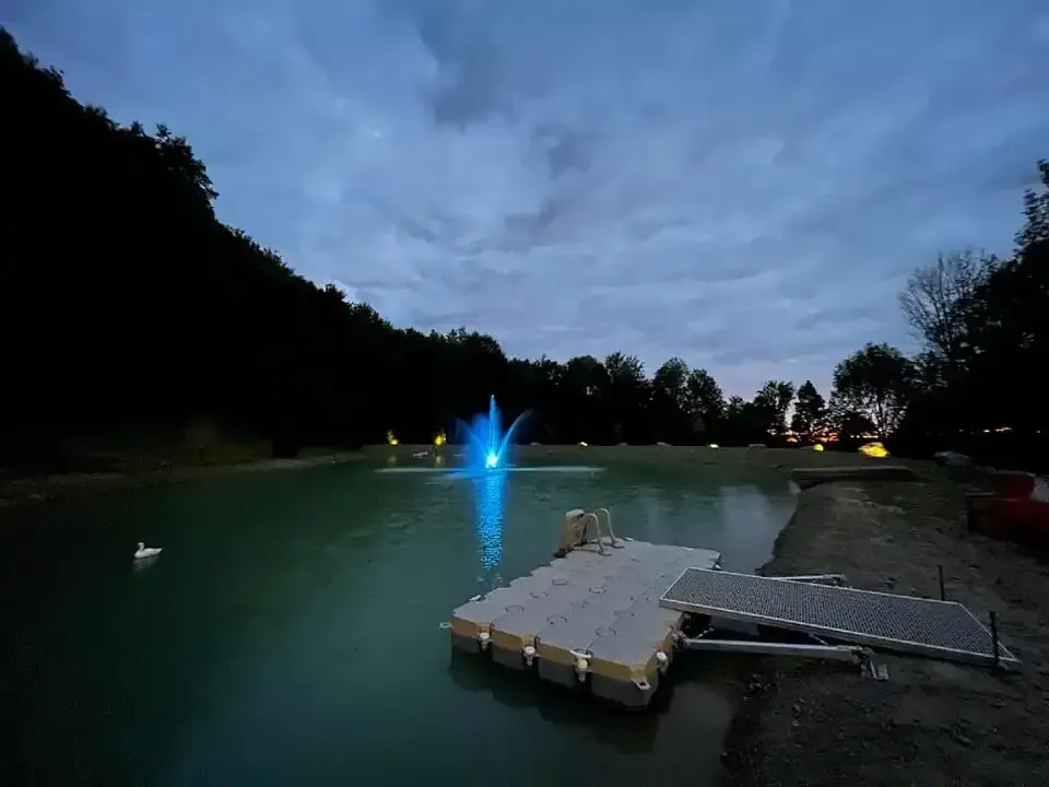 Dusk at a lakeside dock with blue fountain light, calm water, and trees silhouetted along the shore
