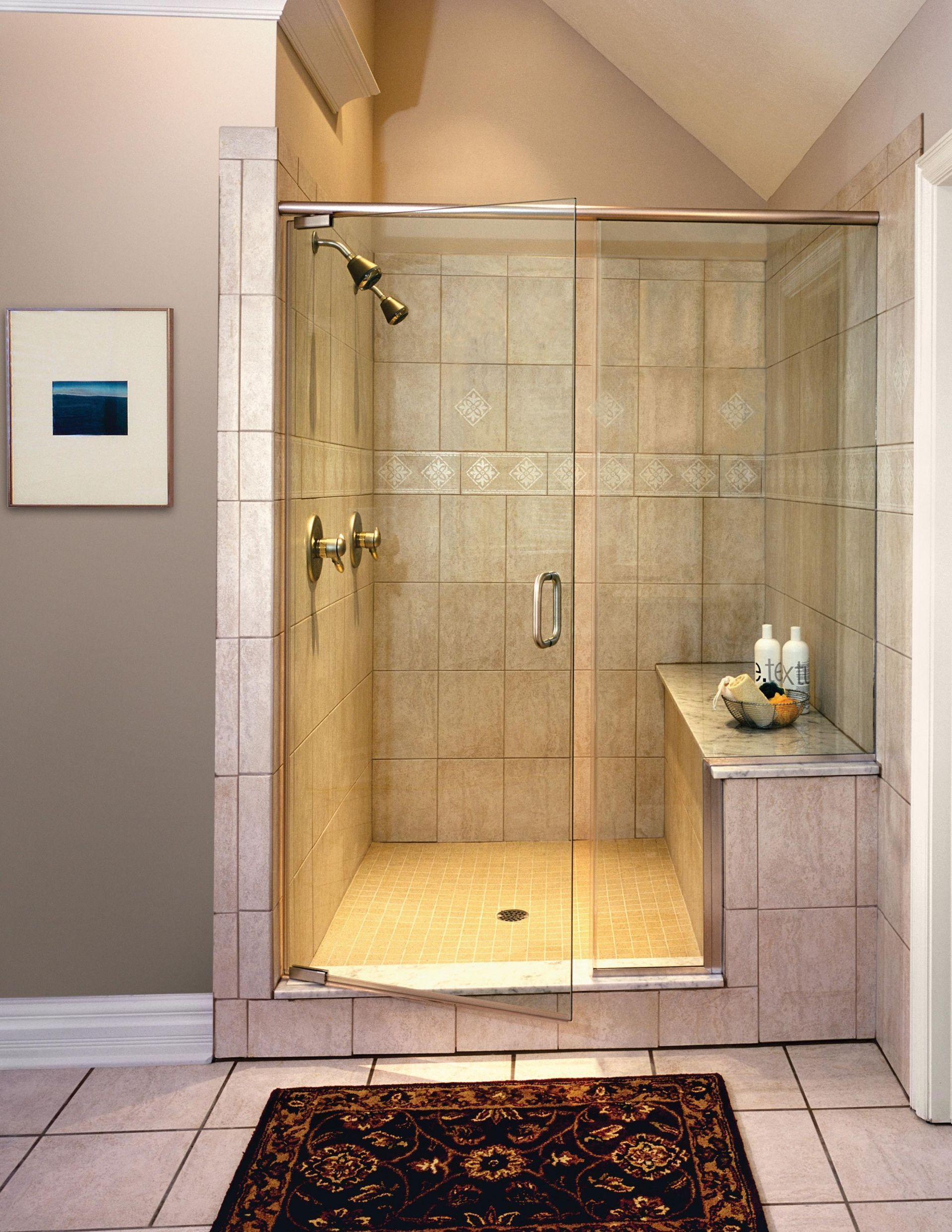 A walk-in shower with beige tile, a built-in stone bench, glass door, and a brass showerhead and handle.