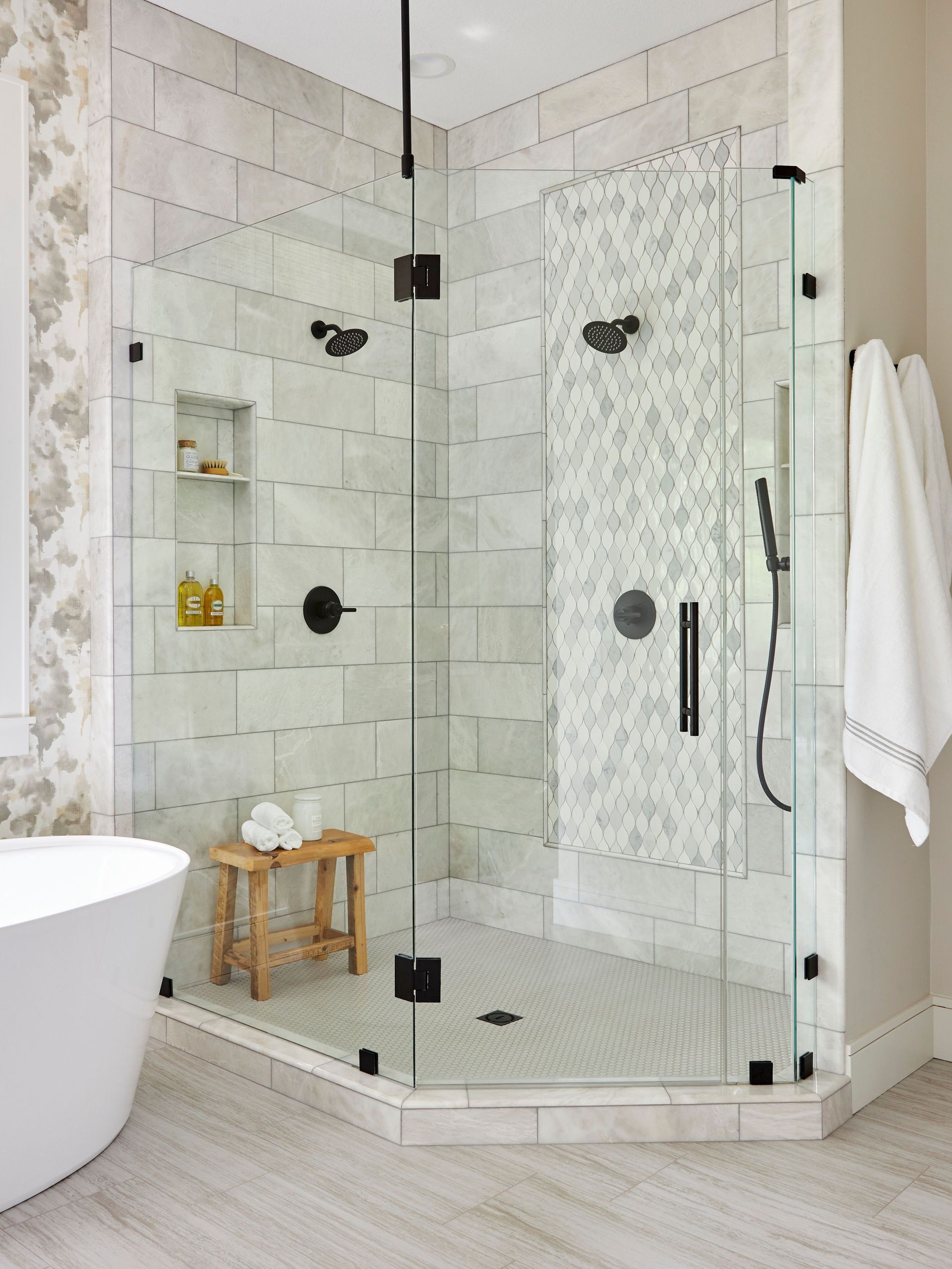 A corner glass shower with light-colored tile walls, a small wooden bench, and matte black fixtures in a modern bathroom.