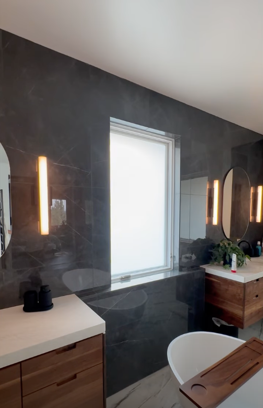 Modern bathroom vanity with dark tiled walls, illuminated mirrors, sink, and a window.