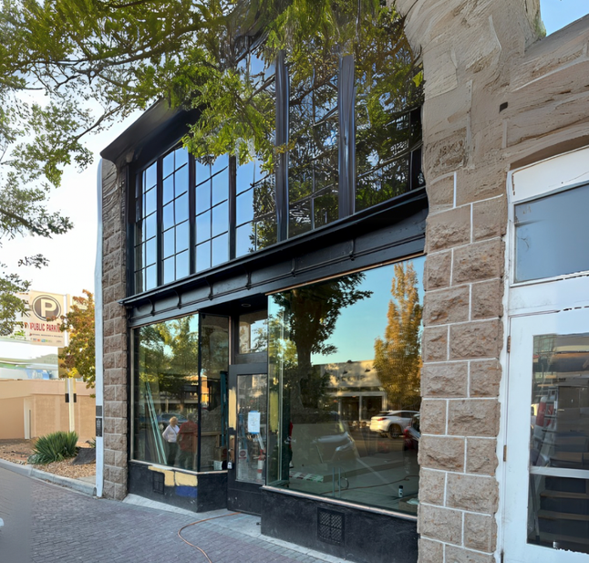 A storefront with large glass windows and black metal framing set into a rough-hewn stone building on a city street.