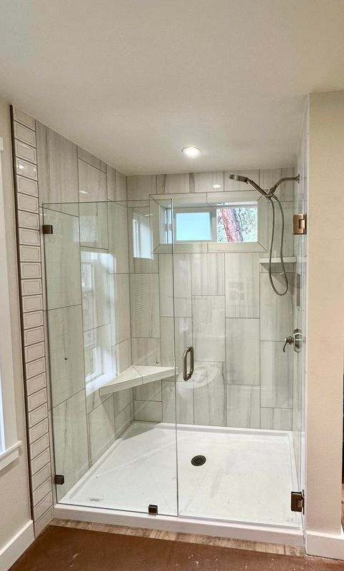 A modern tiled walk-in shower with a glass door, built-in corner seat, handheld showerhead, and a white shower floor.