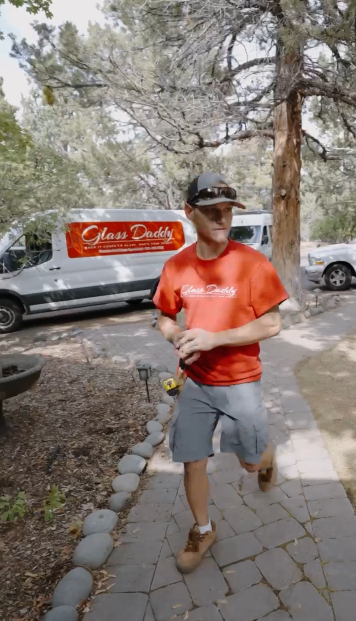 Man in a red shirt walking on a stone path beside a white van in a wooded area