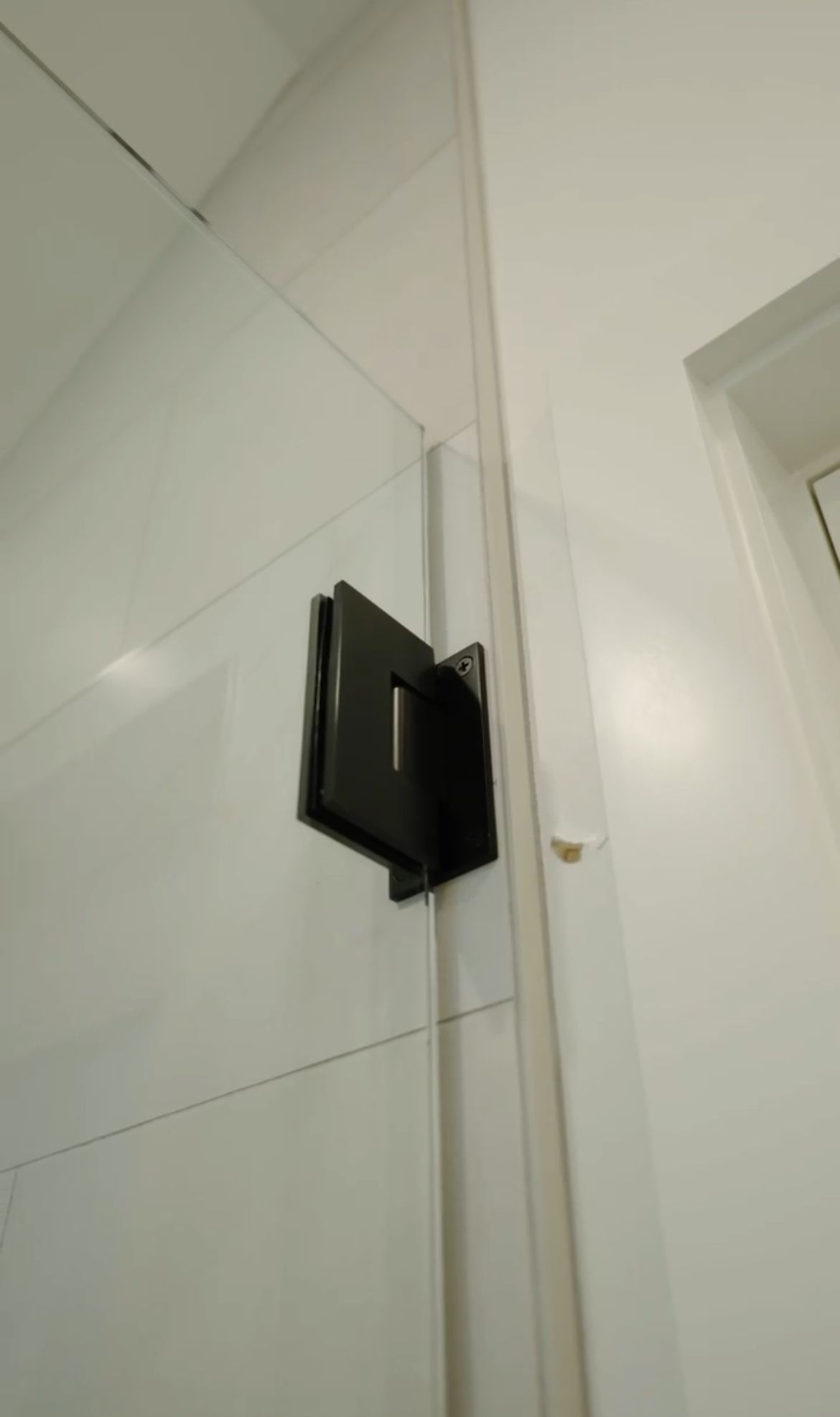 A black metal hinge attaches a glass shower door to a white tiled wall in a bathroom.