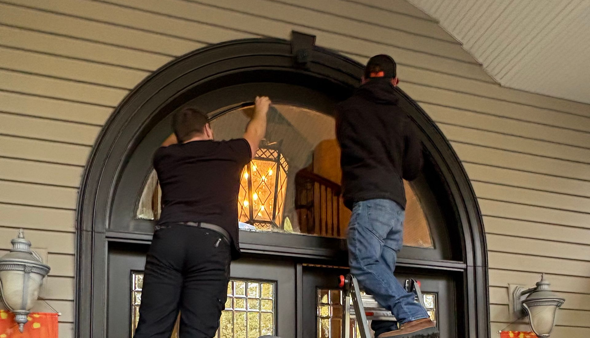 Two people stand on ladders, installing a hanging lantern light fixture inside an arched entryway above double doors.
