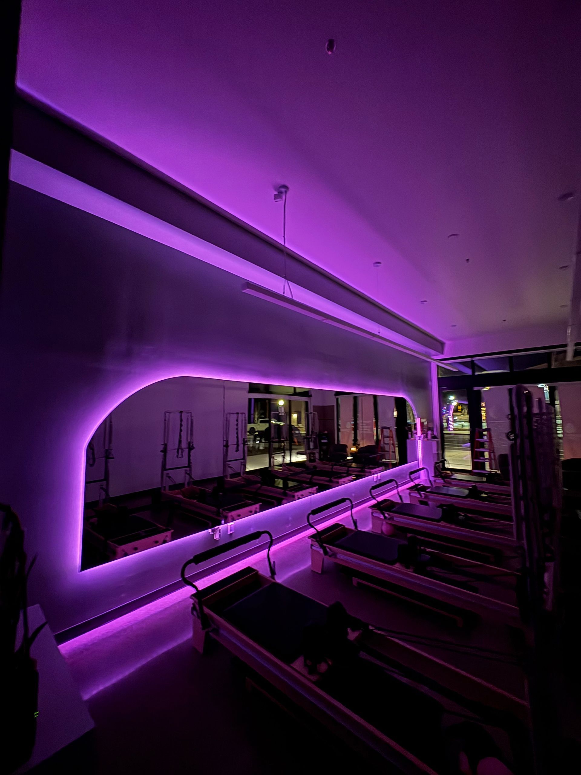 A dimly lit Pilates studio featuring several reformers arranged in rows, illuminated by vibrant purple LED strip lighting.