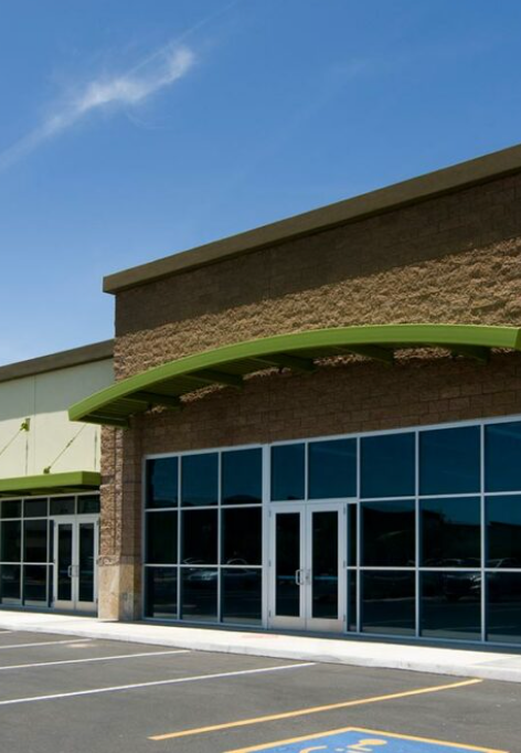 A tan brick commercial building with green awnings and large glass windows and doors under a blue sky.