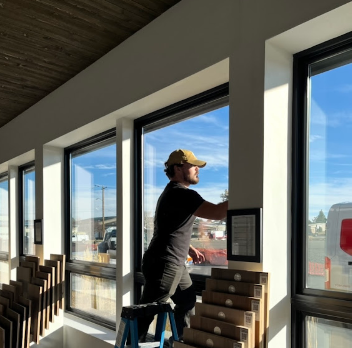 A person in a dark t-shirt and cap stands on a blue stepladder, installing or cleaning large windows in a sunlit room.