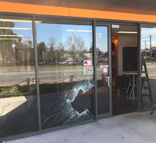 A ground-level view of a business storefront with a large, shattered glass panel next to an open entrance door.