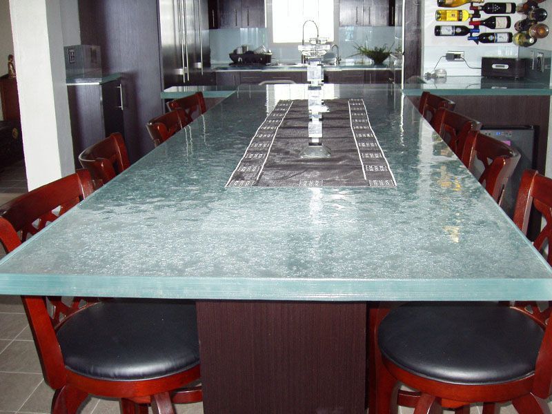 A long, rectangular table with a textured glass top and dark wood base, surrounded by matching chairs in a modern kitchen.