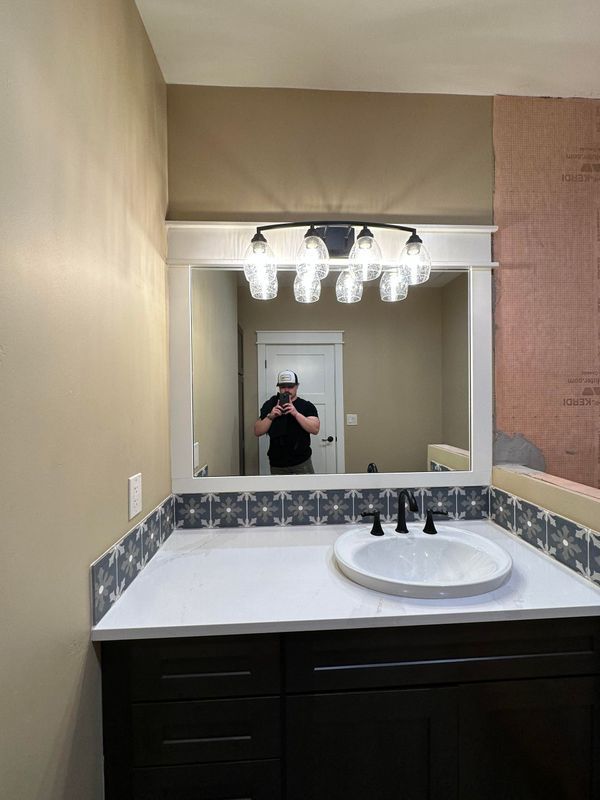 A bathroom vanity with a white countertop, patterned tile backsplash, dark wood cabinets, and an illuminated mirror.
