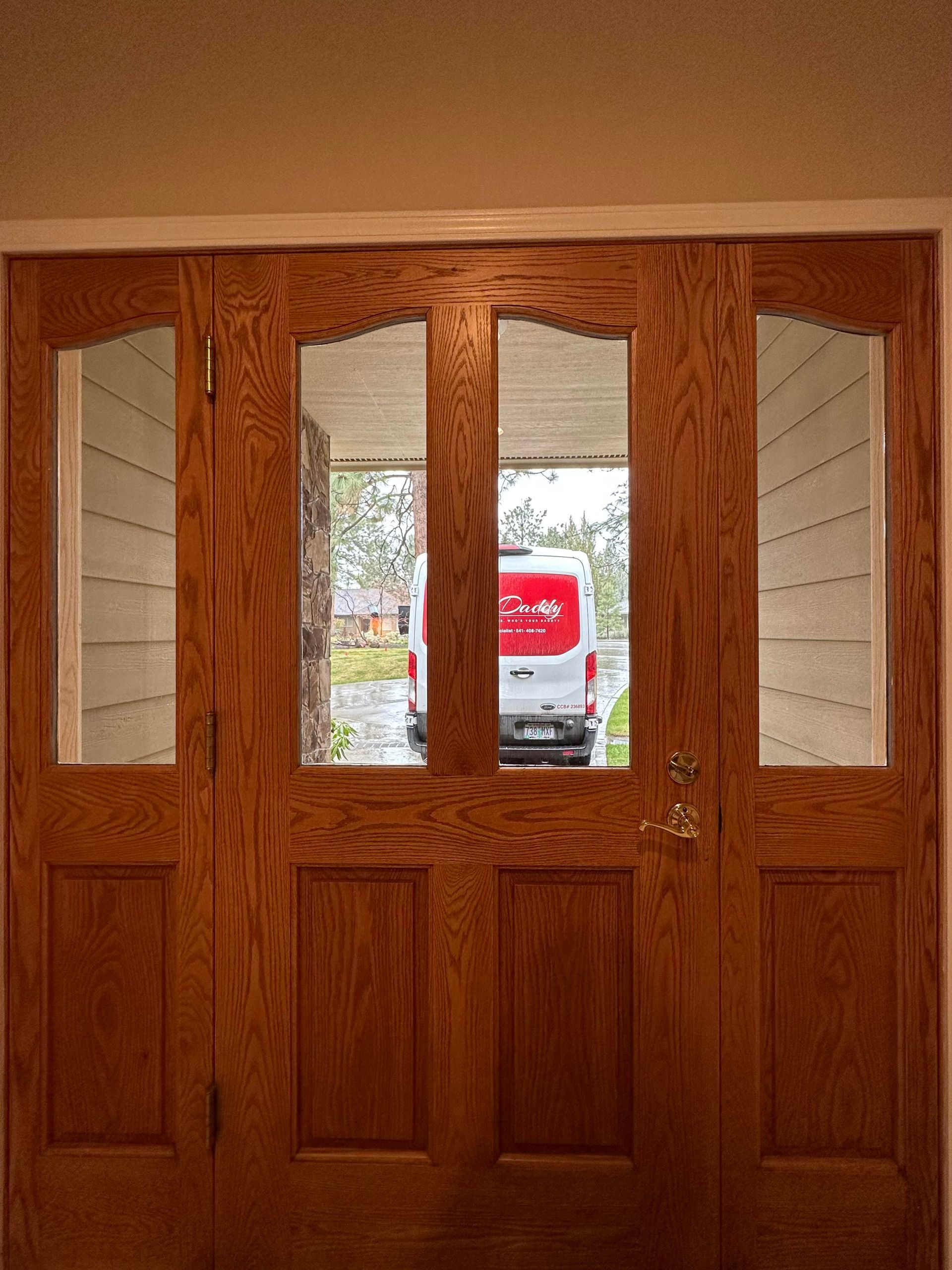 A wooden front door with four glass panels showing a white van parked in the driveway outside.