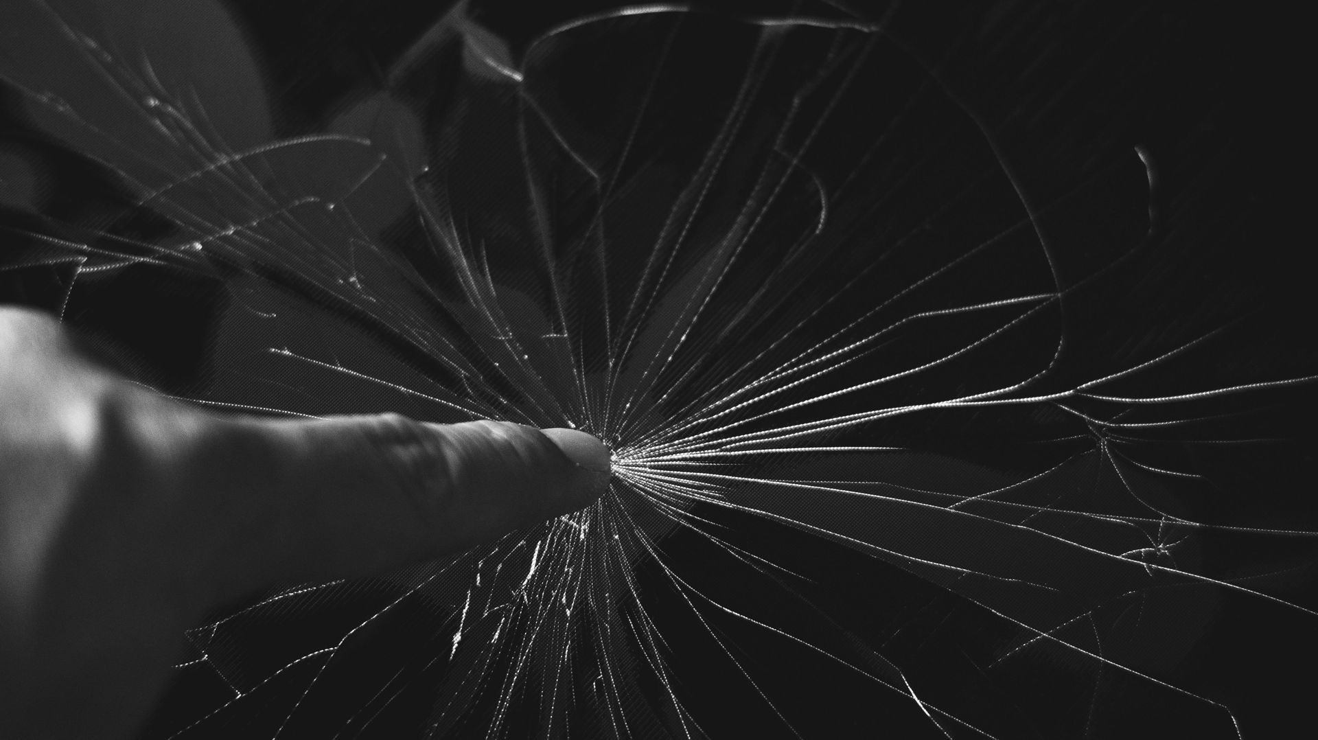 A hand touches a cracked glass surface, centered on the point of impact where radiating lines spread outward.