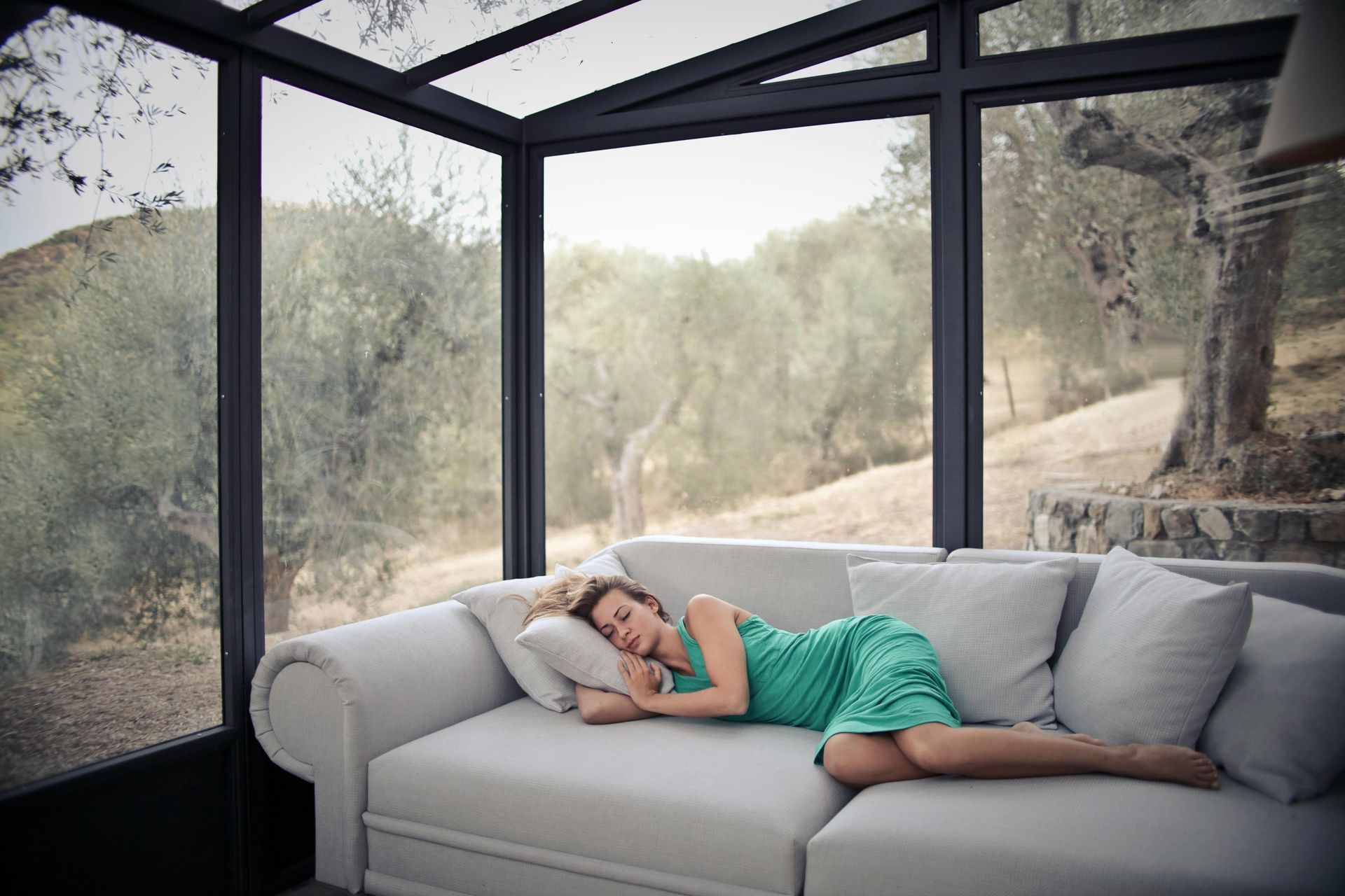 A person in a green dress sleeps on a light-colored sofa inside a glass-walled sunroom overlooking an outdoor landscape.