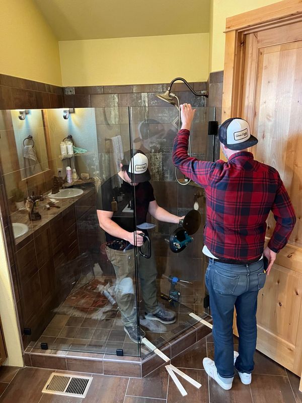 Two professionals in a bathroom install a large glass shower panel.