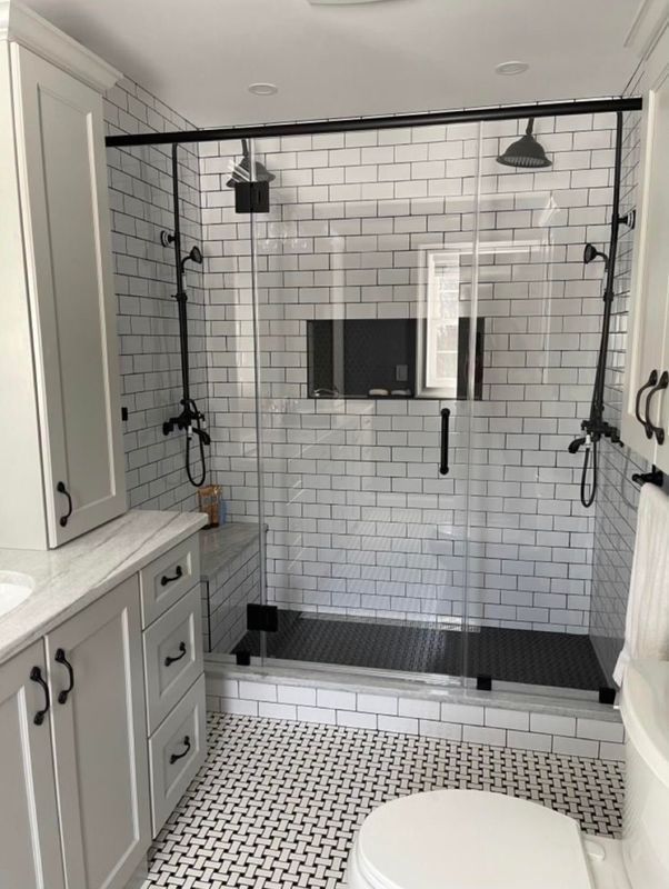 A bathroom featuring a white vanity, a walk-in shower with white subway tile, black hardware, and a patterned tiled floor.