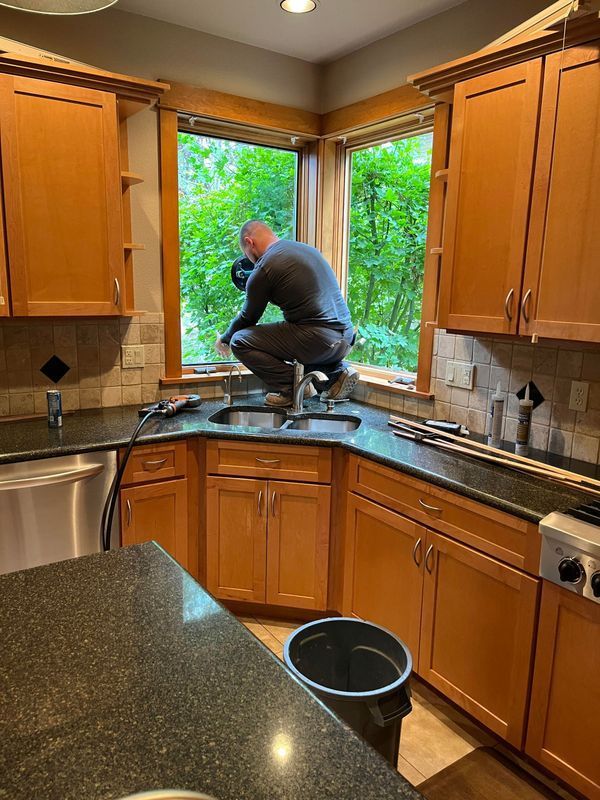 A person crouches on a kitchen sink counter to work on a corner window overlooking trees.