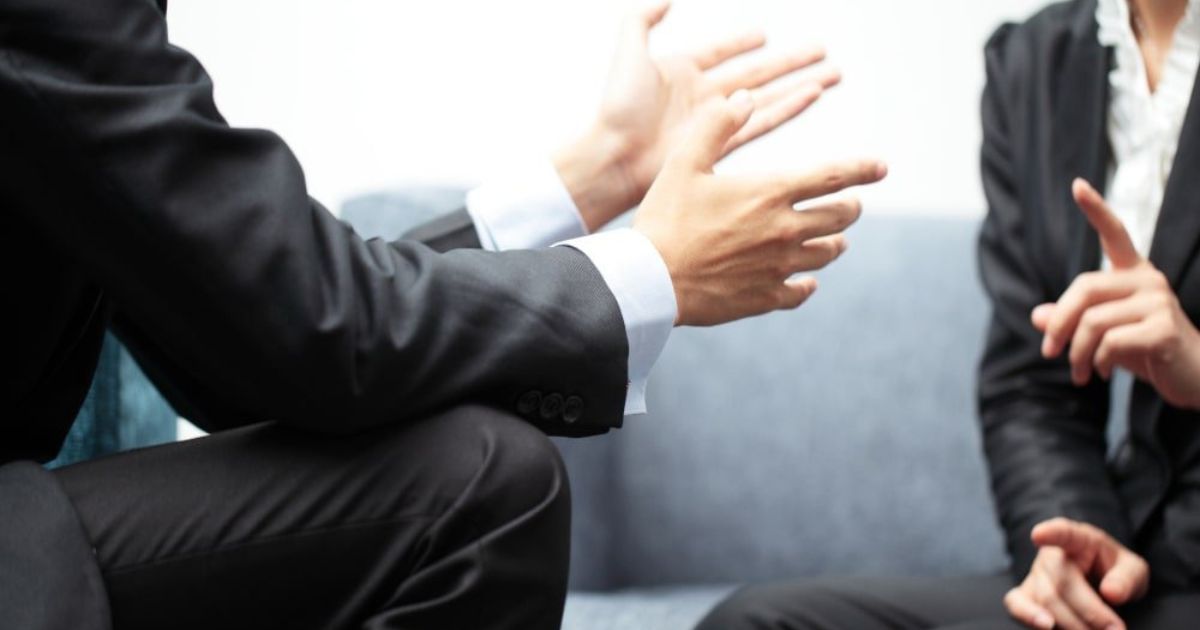 Two people in suits gesturing during a business meeting on a gray couch