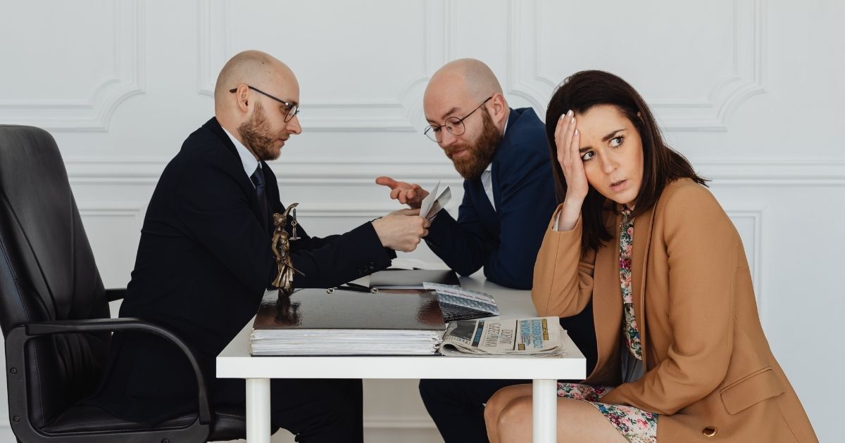 Two men and a woman at a table, money being passed. The woman looks stressed. White room.