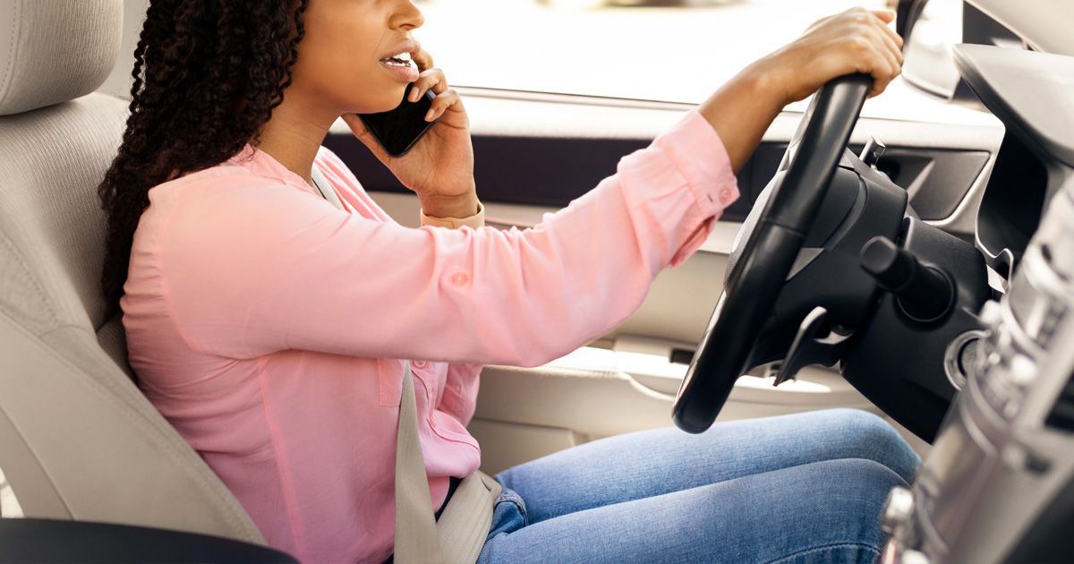 Woman driving a car while talking on a cell phone; wearing pink shirt and blue jeans.