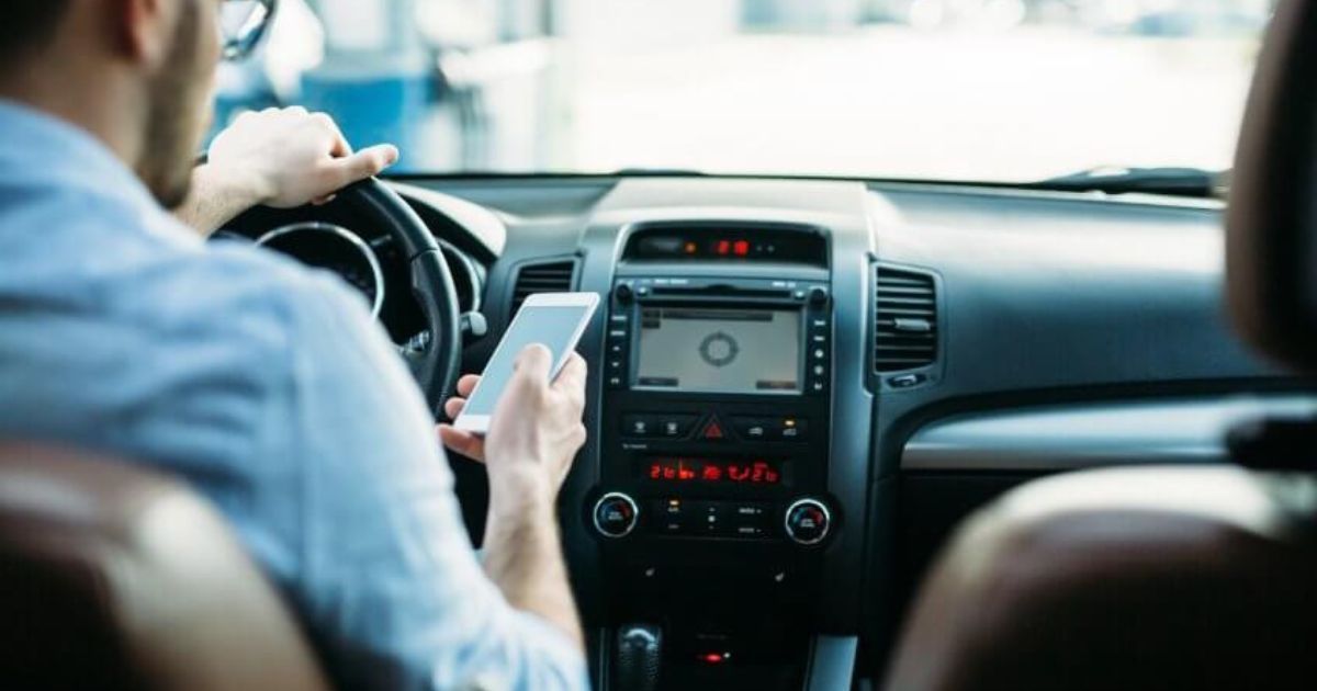 Driver using a smartphone while seated in a car, with the dashboard and steering wheel visible.