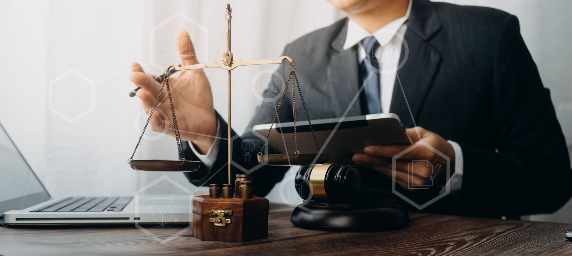 Person in suit holds scales and tablet, near laptop and gavel on a desk. White digital overlay.