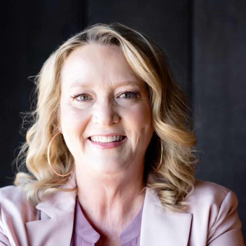 A smiling woman with shoulder-length blonde hair, wearing a light pink blazer, posed against a dark background.