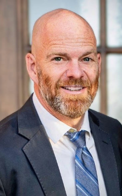 A professional headshot of a smiling person with a beard, wearing a dark suit, light shirt, and blue-striped tie.