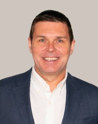 A smiling person with short dark hair, wearing a white collared shirt under a navy blue blazer, against a light gray background.