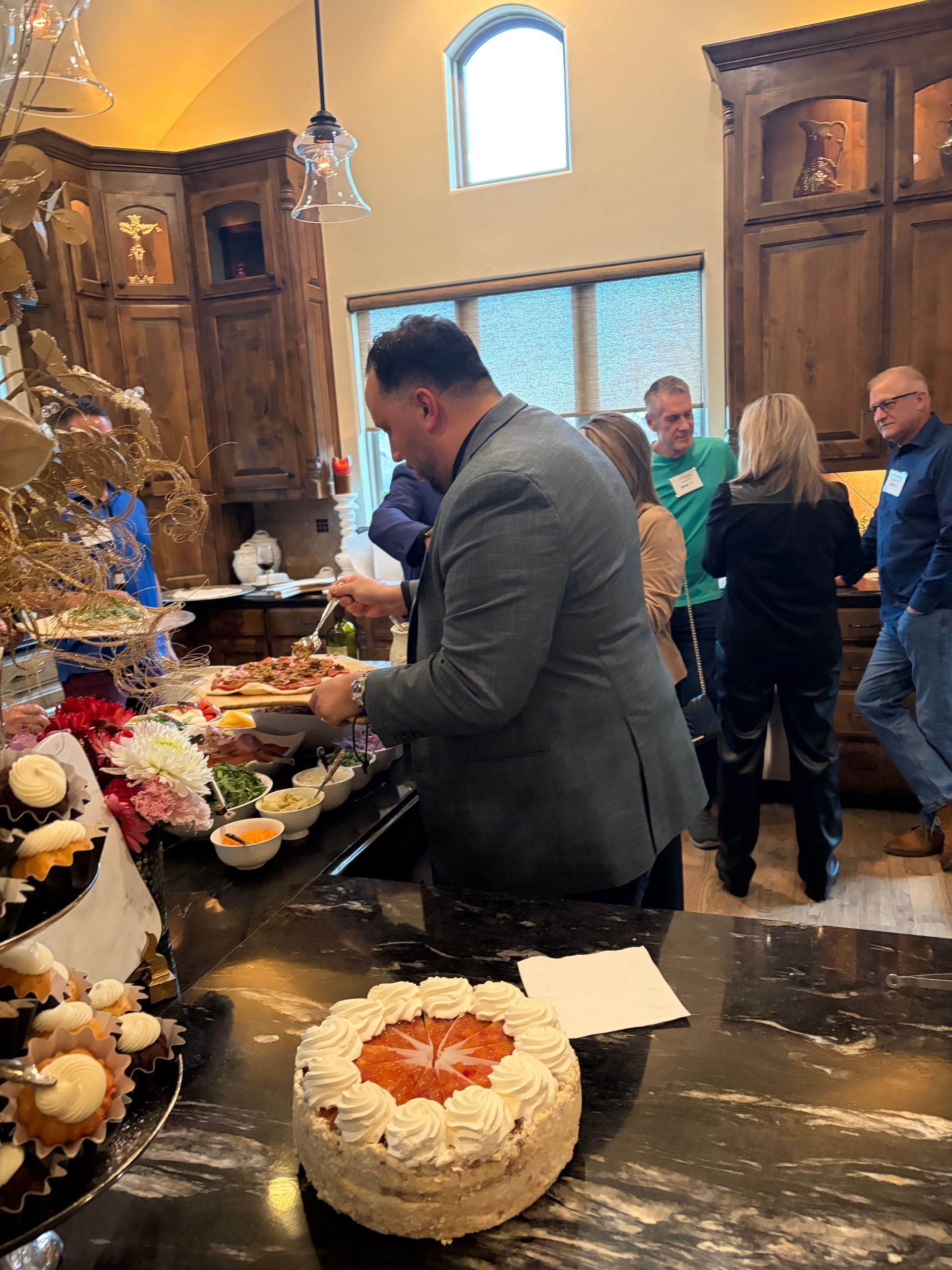 A group of people gathering around a food buffet in a kitchen, with a person serving food from a platter.