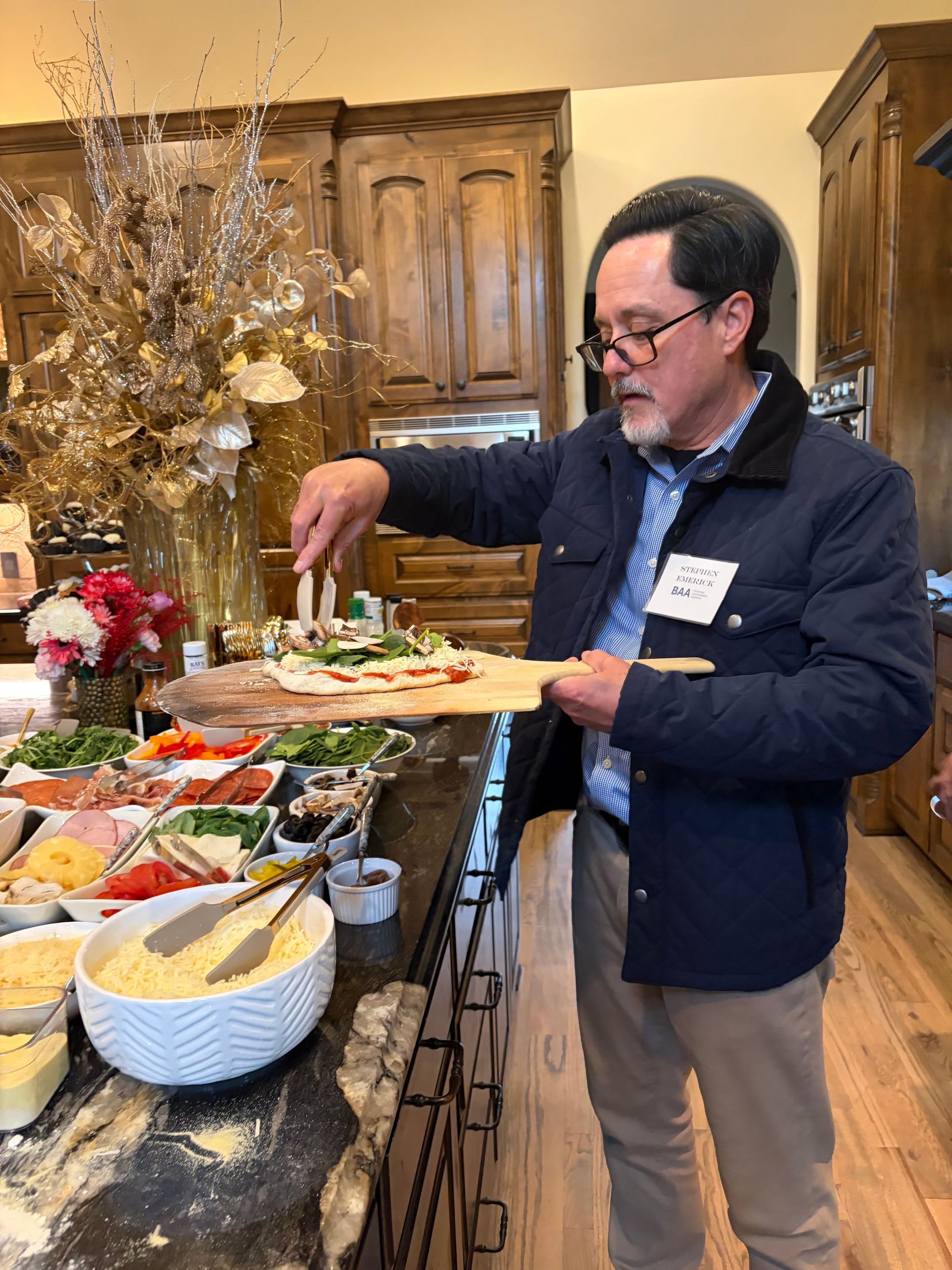 A person in a blue jacket uses tongs to add toppings to a pizza on a wooden peel at a buffet spread in a kitchen.