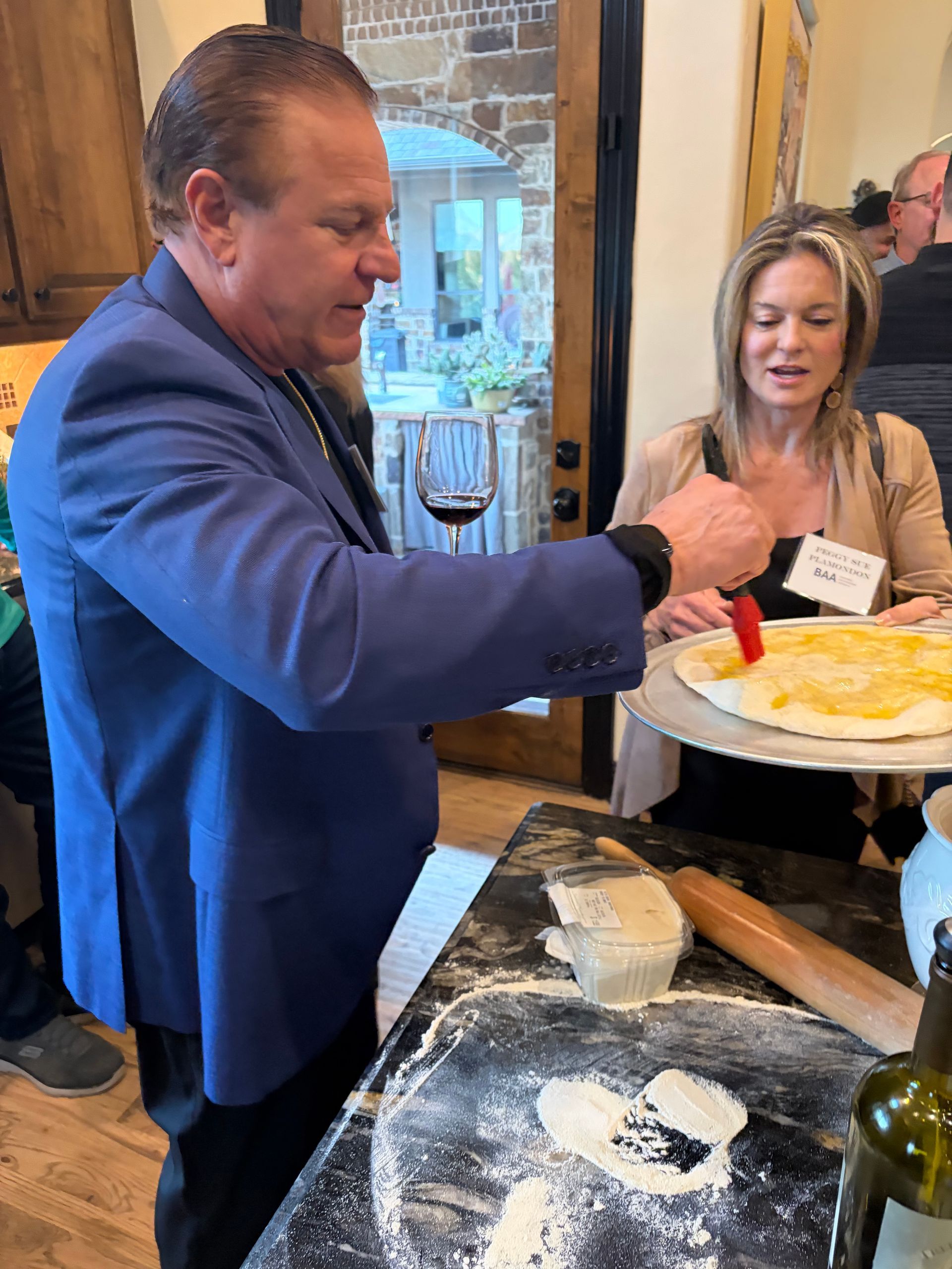 A man in a blue blazer uses a brush to spread oil on pizza dough while a woman watches in a home kitchen setting.