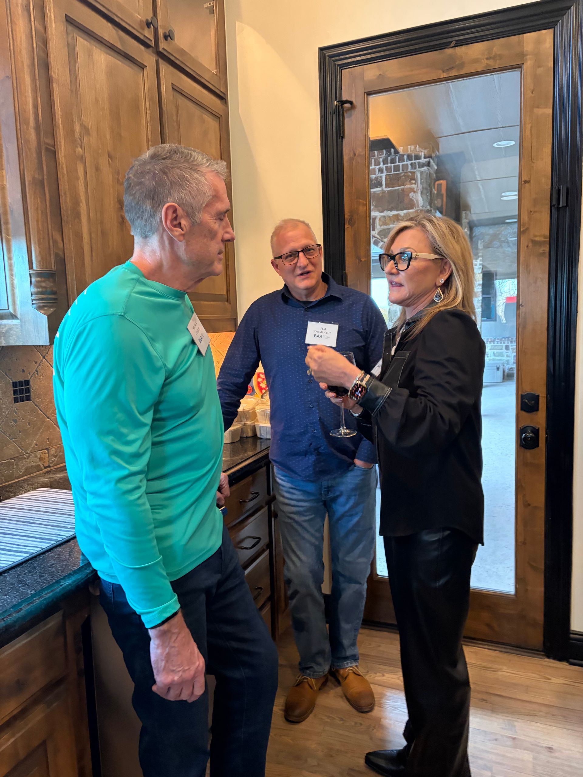 Three people in a kitchen stand in a group, engaged in conversation while holding glasses at a social gathering.