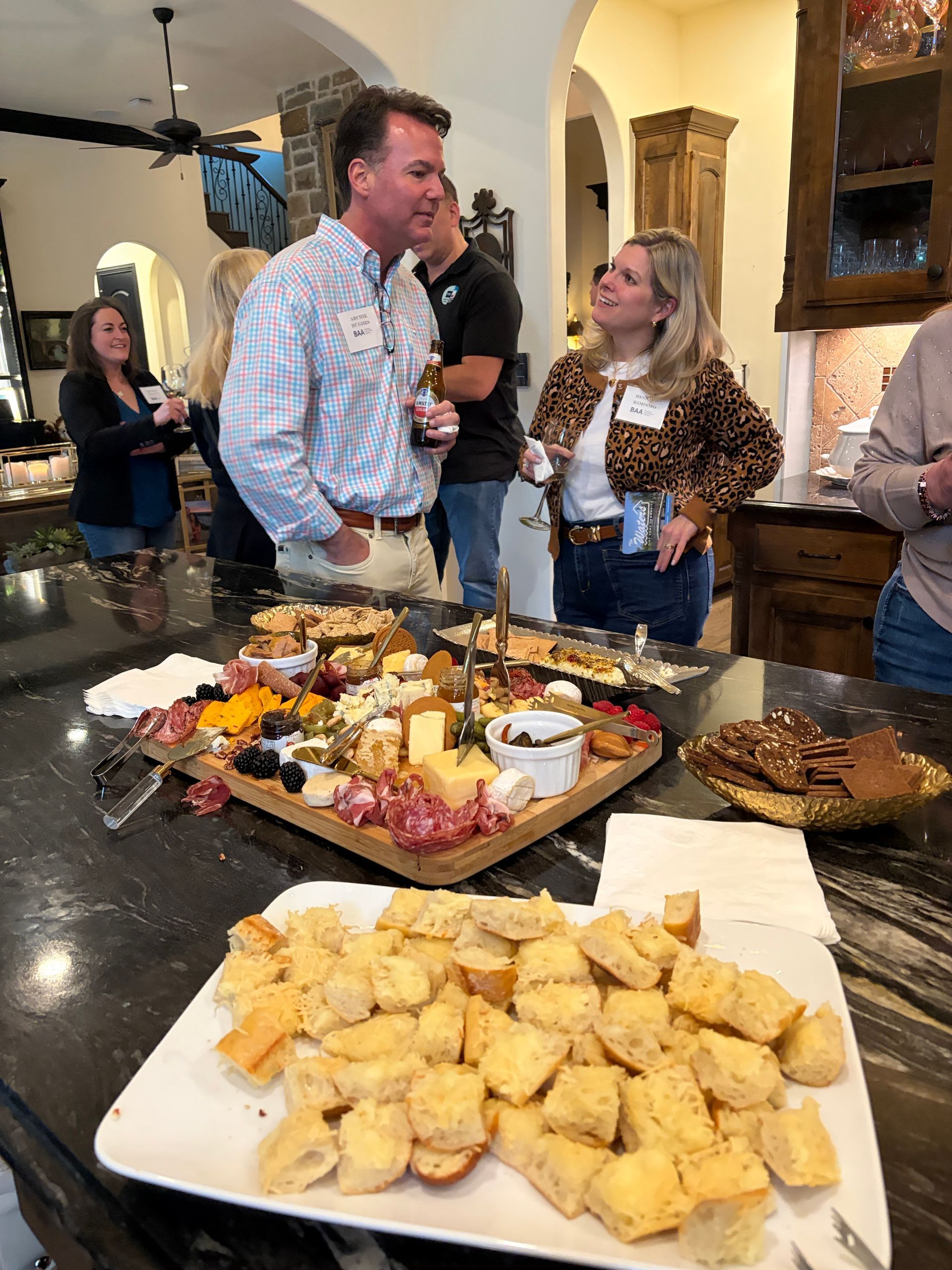 People socialize around a kitchen island filled with charcuterie boards and trays of bread in a home setting.