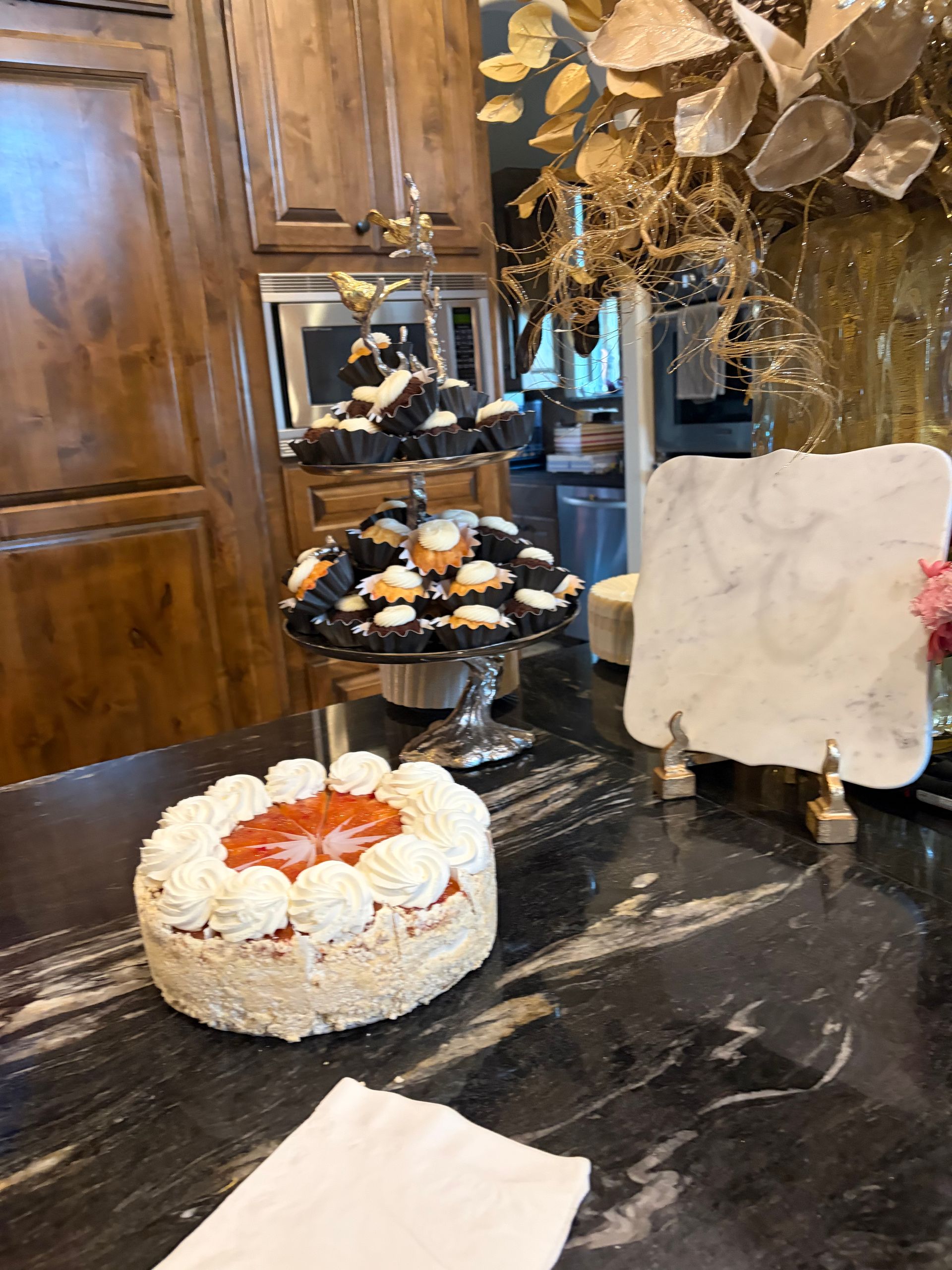 A cake topped with frosting swirls and fruit sits on a dark counter near a tiered dessert stand and a marble tray.