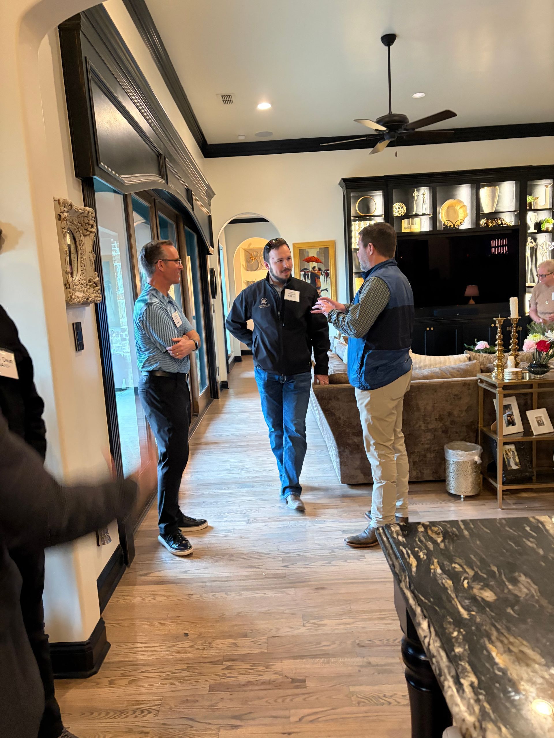 Three people stand conversing in a brightly lit, elegant room featuring hardwood floors and a dark-toned wall display.