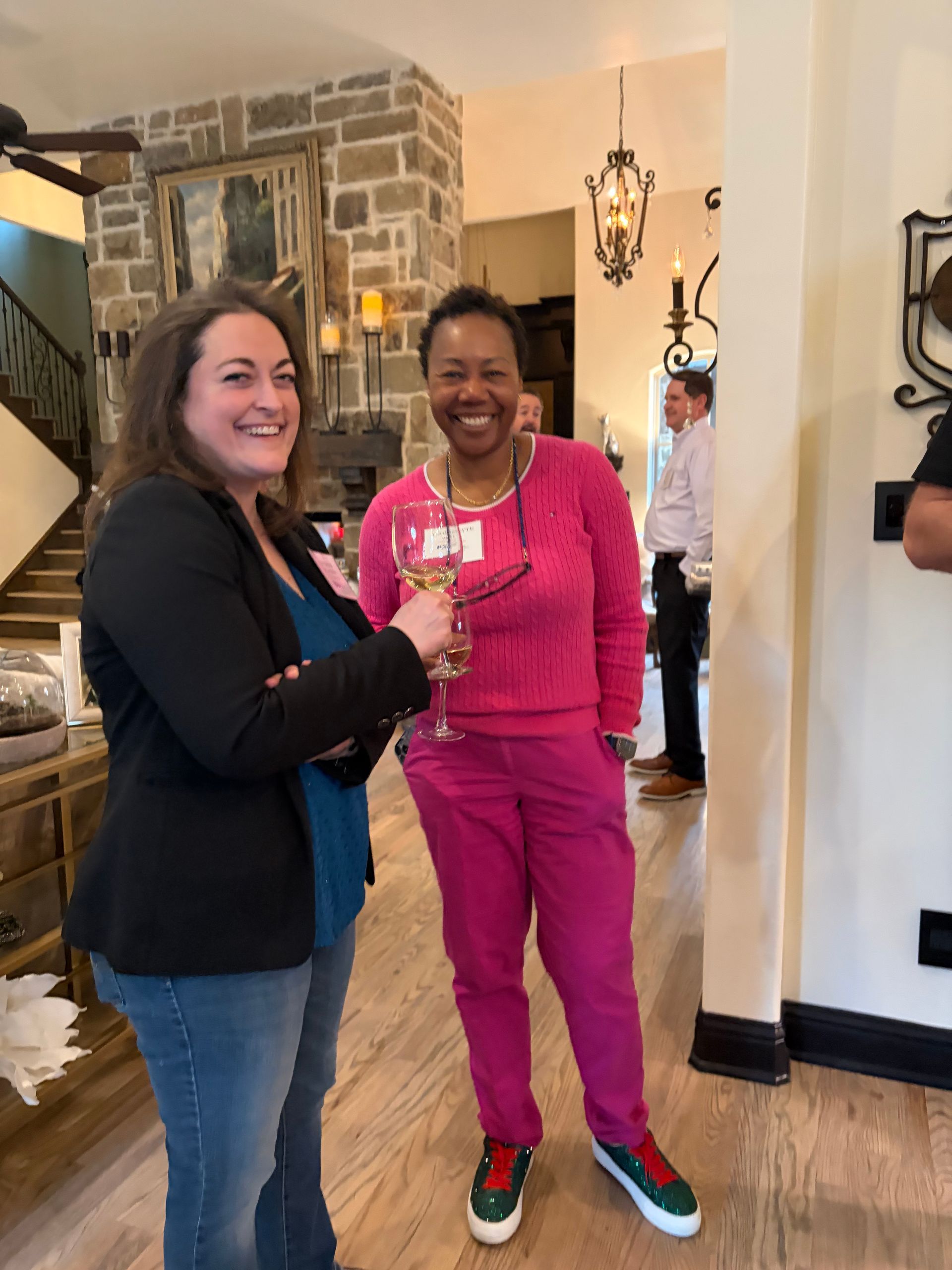 Two women smiling and holding drinks in a well-lit home interior with a stone fireplace and staircase in the background.
