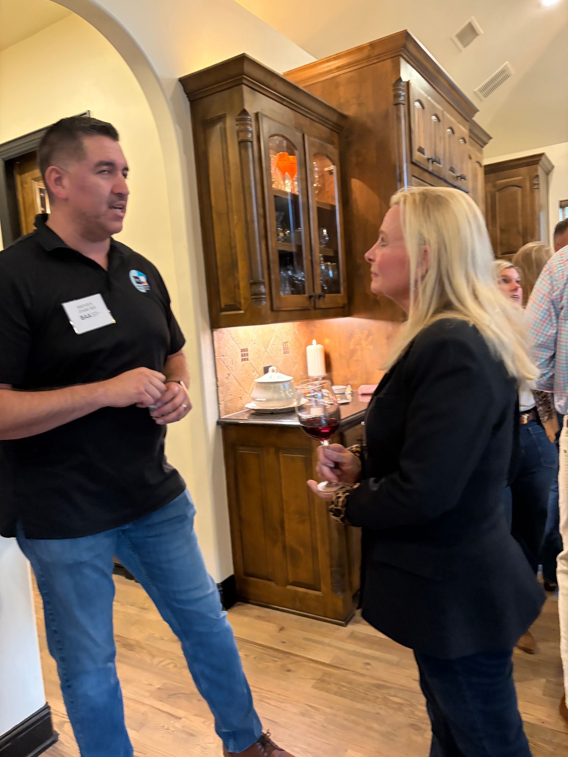 Two people talking in a kitchen; one in a black polo and jeans, the other in a black blazer holding a glass of wine.