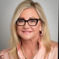 A smiling professional woman with blonde hair wearing dark-rimmed glasses, a pink collared shirt, and a necklace.