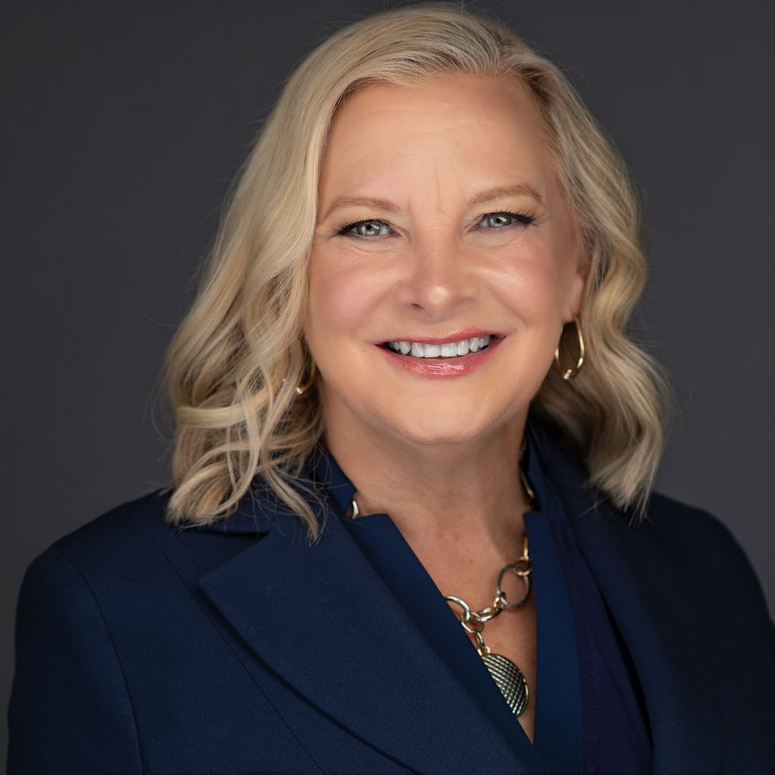 Professional portrait of a blonde individual smiling, wearing a navy blue blazer and a gold link necklace against a grey.