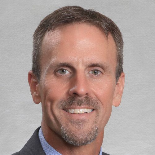 Headshot of a person with short brown hair and a goatee, wearing a dark suit and light blue shirt against a gray backdrop.