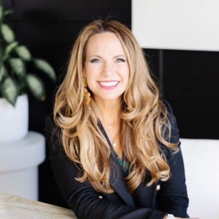 A smiling professional with long wavy hair, wearing a dark blazer and gold earrings, sitting in a modern office space.