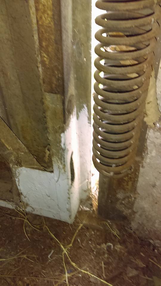Close-up of a weathered, metal spring and wooden door frame with white paint and debris.