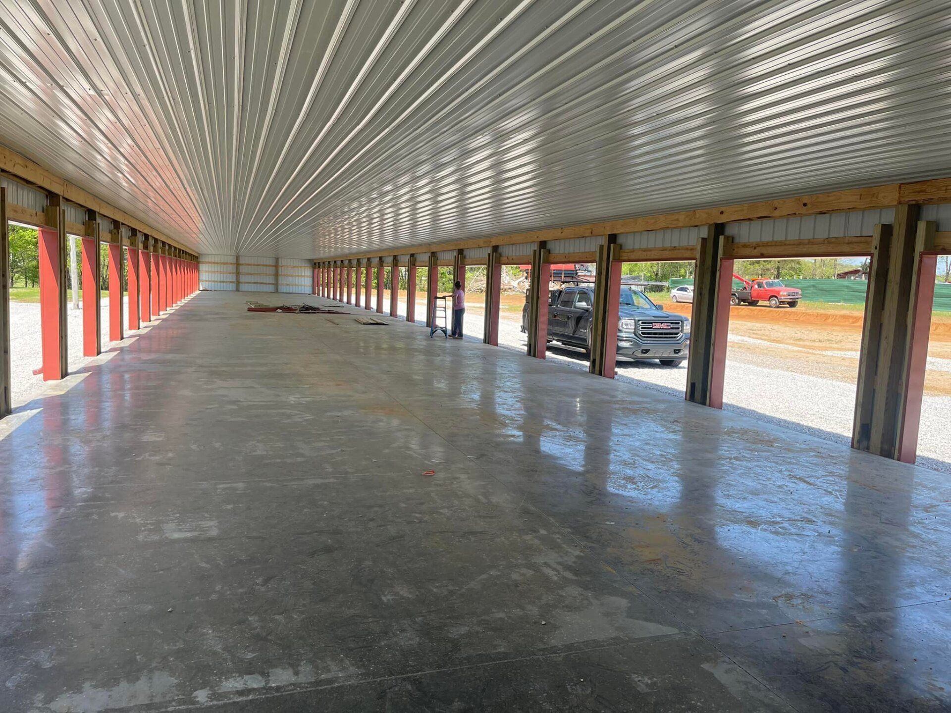 Interior of a large, open metal building with a concrete floor, red support beams, and a corrugated metal ceiling.